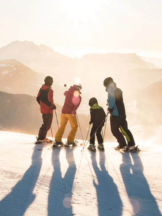 Une famille composée de deux enfants se tenant sur des skis en haut d'une piste contemple le coucher de soleil avec vue sur la chaîne des Aravis. L'un a un ensemble de ski bleu, noir et vert, un autre rouge et jaune, un autre vert et noir et le dernier rouge et noir. L'un des skieurs au milieu pointe avec son bâton gauche le sommet d'une montagne. L'atmosphère est très lunaire avec une lumière blanche/dorée. C'est une belle fin de journée de ski.
