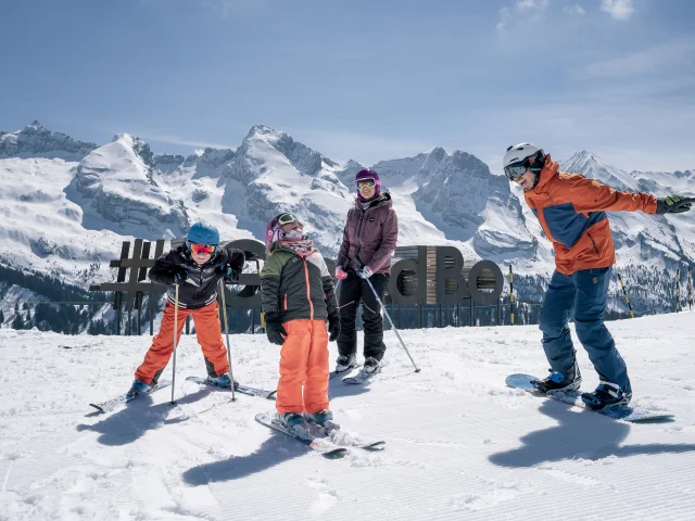 Une famille joyeusement réunie sur les pistes de ski, vêtue de combinaisons colorées et de casques, affiche des sourires éclatants alors qu'ils se tiennent côte à côte, skis et snowboard en main, prêts à dévaler les pentes enneigées. Nous apercevons en arrière-plan la sculpture #monGrandBo et la chaîne des Aravis d'un blanc éclatant. Cette image évoque non seulement la joie de skier ensemble, mais aussi les précieux moments de partage et de complicité en famille, faisant de chaque descente une expérience mémorable.