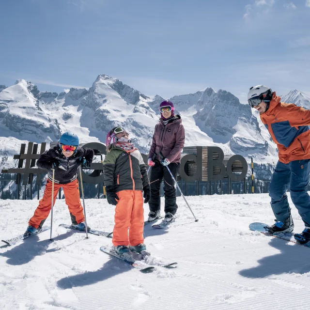 Une famille joyeusement réunie sur les pistes de ski, vêtue de combinaisons colorées et de casques, affiche des sourires éclatants alors qu'ils se tiennent côte à côte, skis et snowboard en main, prêts à dévaler les pentes enneigées. Nous apercevons en arrière-plan la sculpture #monGrandBo et la chaîne des Aravis d'un blanc éclatant. Cette image évoque non seulement la joie de skier ensemble, mais aussi les précieux moments de partage et de complicité en famille, faisant de chaque descente une expérience mémorable.