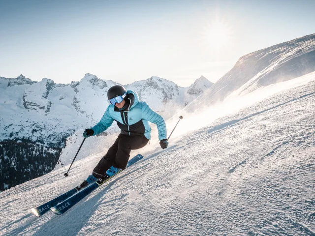 Skier in a light blue and black ski suit skiing down a slope with a view of the Aravis mountain range and a clear sky in the background
