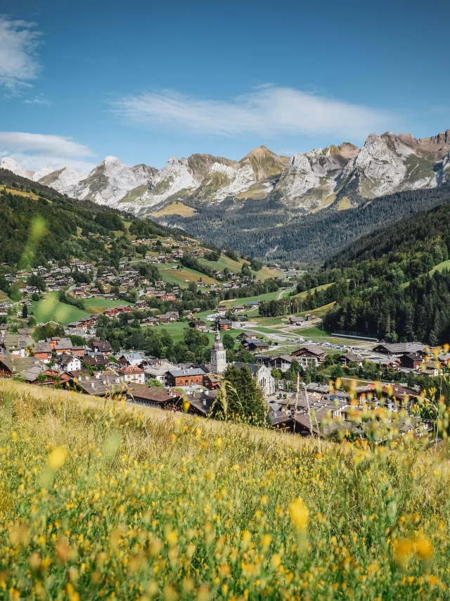 Vue du Village en été du Grand-Bornand avec des fleurs jaunes floues en premier plan. Belle vue sur la chaîne des Aravis, dans une nature verdoyante.