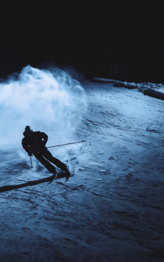 Skieur en pleine descente nocturne, illuminé par un projecteur, laissant une traînée de poudreuse dans la nuit.