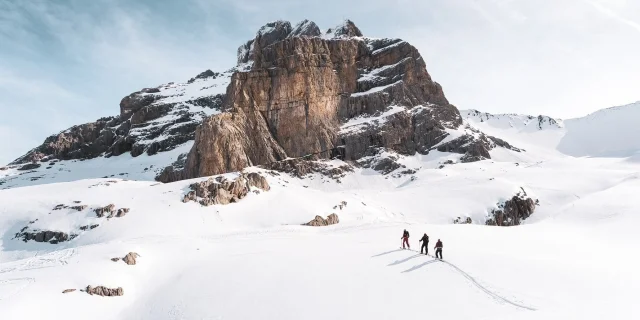 Three skiers advance across a vast expanse of snow, with a rocky mountain in the background, under a pale blue sky.