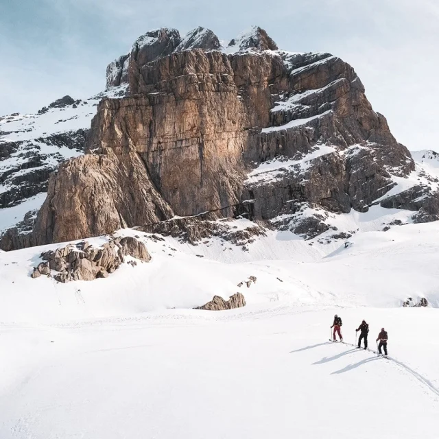 Trois skieurs avancent sur une vaste étendue enneigée, avec une montagne rocheuse en arrière-plan, sous un ciel bleu pâle.