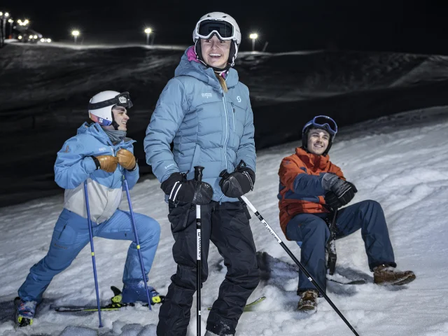 Trois skieurs habillés chaudement profitent d'une session de ski nocturne sur une piste enneigée éclairée par des projecteurs, avec un fond de montagne plongé dans l'obscurité.