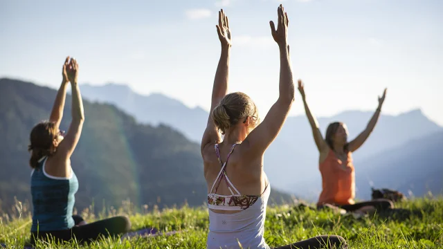 A group of 3 people sitting on the grass raise their hands to the sky in a yoga position, facing a mountainous landscape under a blue sky.