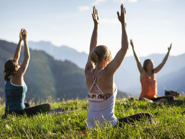 Un groupe de 3 personnes assis dans l'herbe lèvent les mains vers le ciel dans une position de yoga, face à eux se trouve un paysage montagneux sous un ciel bleu.