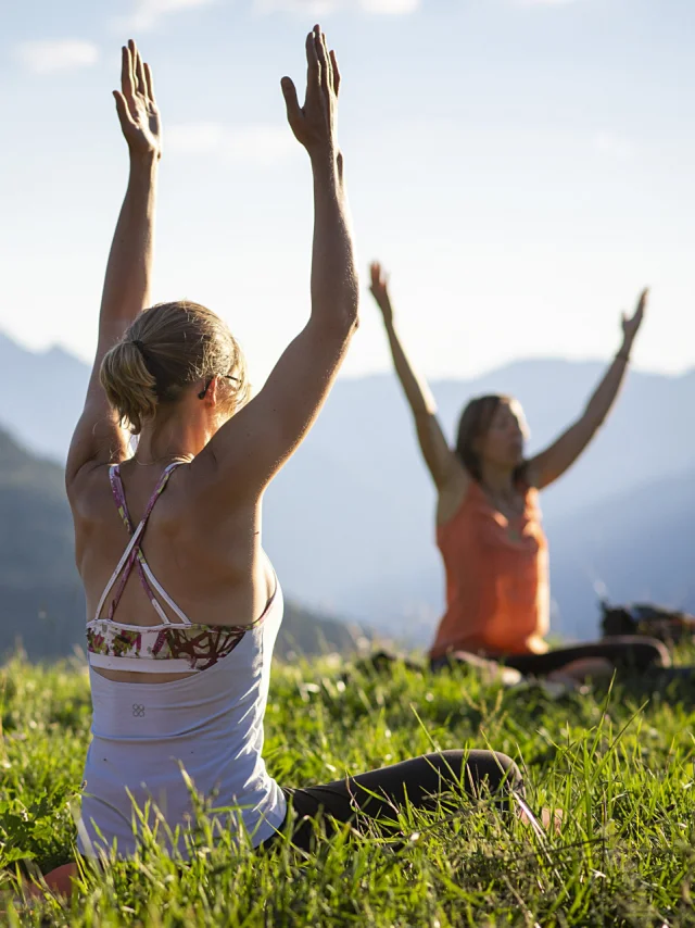 Un groupe de 3 personnes assis dans l'herbe lèvent les mains vers le ciel dans une position de yoga, face à eux se trouve un paysage montagneux sous un ciel bleu.