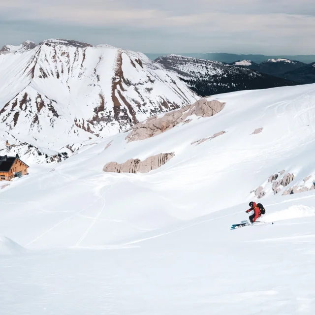Un skieur fait un virage dans une vaste étendue de neige vierge au dessus du refuge de la pointe percée et du col des annes