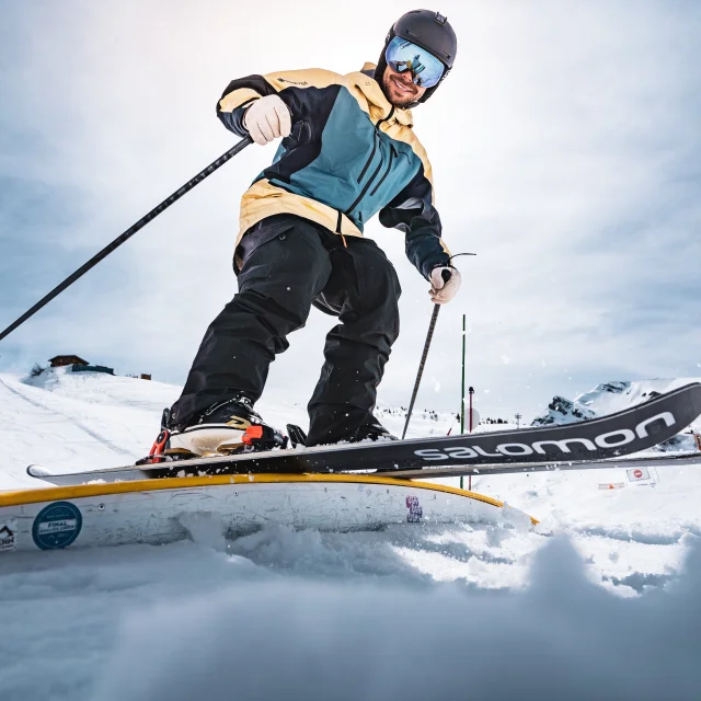 Un skieur freestyle est pris en photo du dessous en train de glisser sur une box avec des skis salomon alors qu'en arrière plan se trouve un paysage de montagnes enneigés