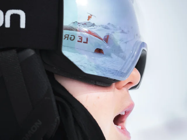 photo d'un visage juvenile prise de profil qui laisse apparaitre un skieur sautant sur l'airbag du snowparkgb dans le reflet de son masque