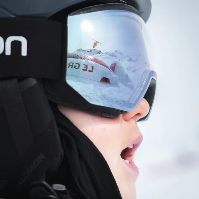 photo d'un visage juvenile prise de profil qui laisse apparaitre un skieur sautant sur l'airbag du snowparkgb dans le reflet de son masque