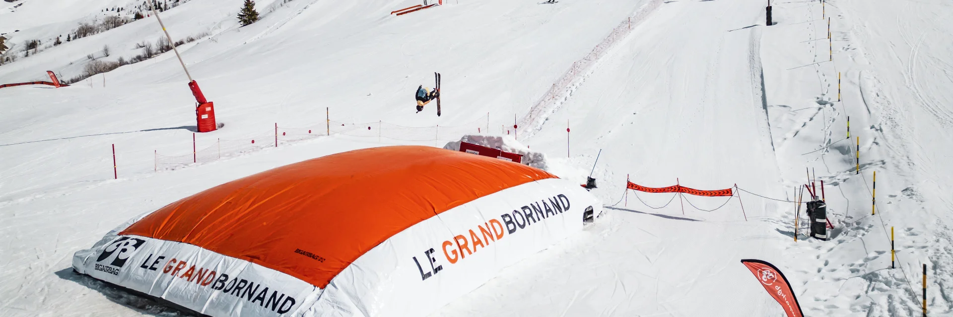 Un skieur effectue une figure en l’air avant d’atterrir sur un grand airbag orange marqué Le Grand-Bornand, dans un décor enneigé et montagneux.