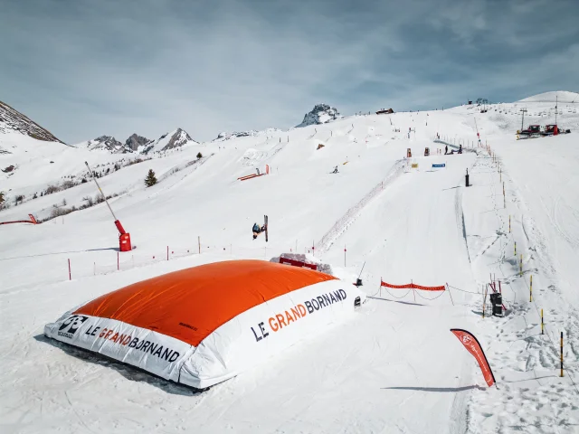 Un skieur effectue une figure en l’air avant d’atterrir sur un grand airbag orange marqué Le Grand-Bornand, dans un décor enneigé et montagneux.