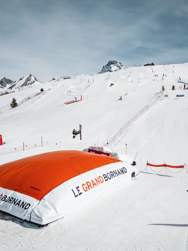 Un skieur effectue une figure en l’air avant d’atterrir sur un grand airbag orange marqué Le Grand-Bornand, dans un décor enneigé et montagneux.