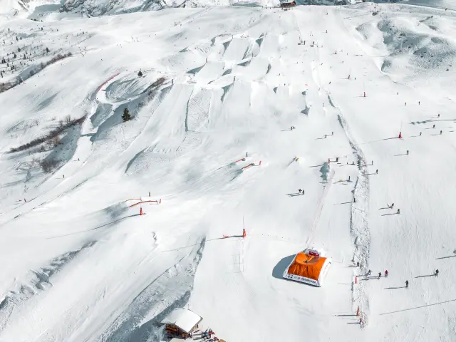 Vue aérienne du snowpark du Grand-Bornand avec ses modules et pistes enneigées, entouré par la chaine des Aravis sous un ciel dégagé.