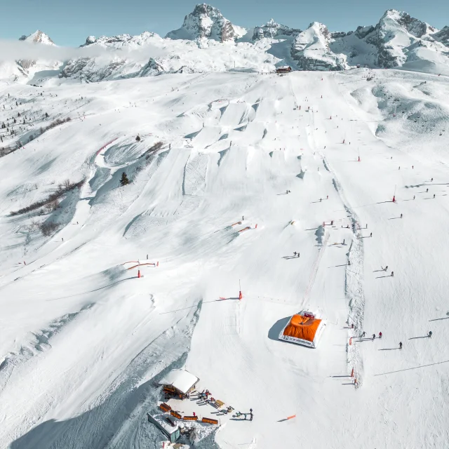 Vue aérienne du snowpark du Grand-Bornand avec ses modules et pistes enneigées, entouré par la chaine des Aravis sous un ciel dégagé.