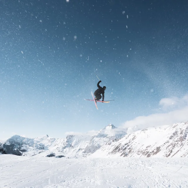 Un skieur freestyle est en l'air et grab ses skis tandis qu'il amène avec lui de la neige qui vole dans les airs et devant lui se trouvent des montagnes enneigées.