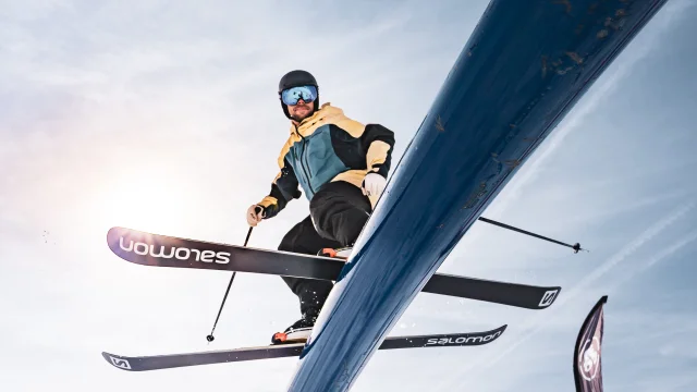 Un skieur freestyle est pris en photo par le dessous pendant qu'il glisse sur un tube en métal avec des pentes einneigés et le soleil derrière lui.