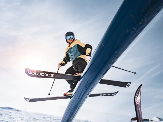 A freestyle skier is photographed from below as he glides down a metal tube with snow-covered slopes and the sun behind him.