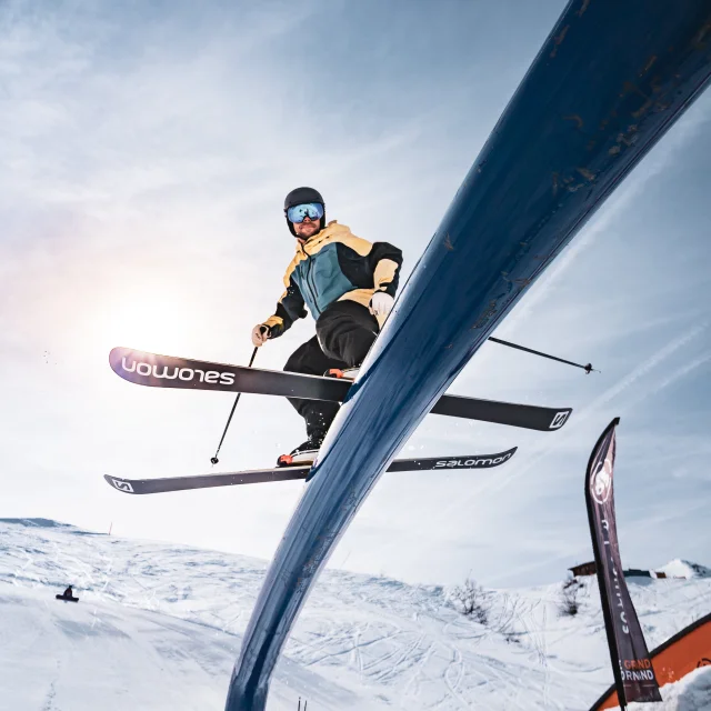 Un skieur freestyle est pris en photo par le dessous pendant qu'il glisse sur un tube en métal avec des pentes einneigés et le soleil derrière lui.