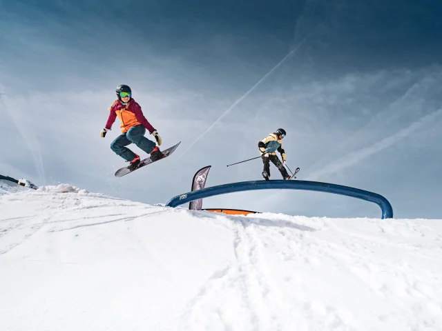 A snowboarder makes a jump while a skier slides on a rainbow rail at the Grand-Bornand snowpark, under clear skies.