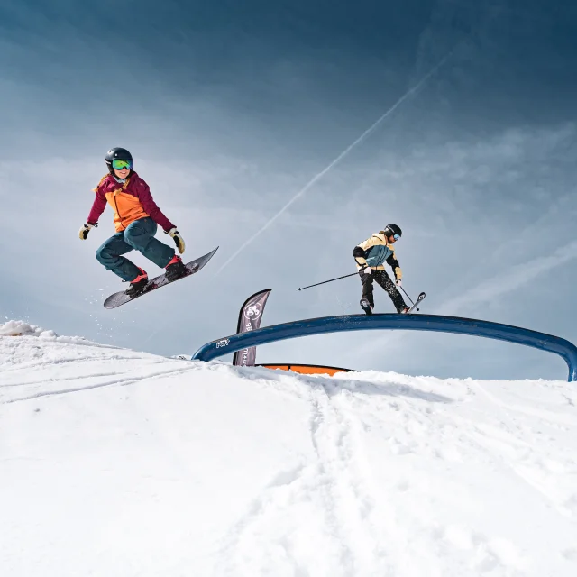 Un snowboarder effectue un saut tandis qu’un skieur slide sur un rainbow rail au snowpark du Grand-Bornand, sous un ciel dégagé.