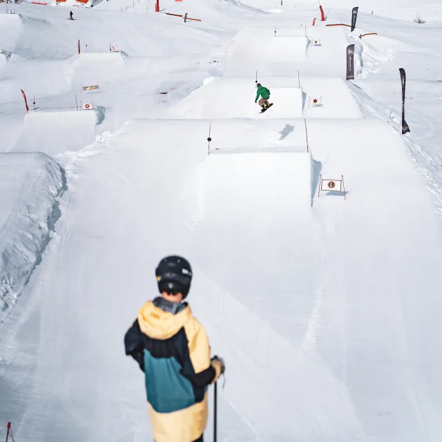 Un skieur de dos au départ de la ligne de sauts de niveau L du snowpark du grand-bornand pendant qu'au loin un snowboarder se trouve dans les airs