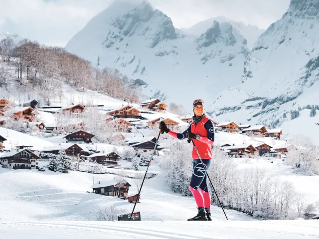 Une skieuse de fond sur une piste de ski nordique avec en arrière plan le village du grand-bornand et la chaine des Aravis enneigées