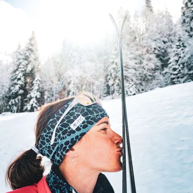 Une skieuse embrasse affectueusement ses skis, exprimant sa passion pour le ski nordique, avec en arrière-plan un paysage enneigé et une forêt givrée.