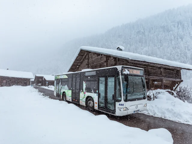 Un bus de transport public stationné sur une route enneigée devant un chalet en bois, avec une forêt de sapins enneigée en arrière-plan.