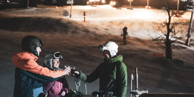 Un groupe de skieurs trinque en terrasse, derrière eux se trouve une piste de ski nocturne éclairée ainsi qu'un télésiège