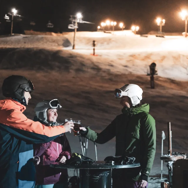 Un groupe de skieurs trinque en terrasse, derrière eux se trouve une piste de ski nocturne éclairée ainsi qu'un télésiège