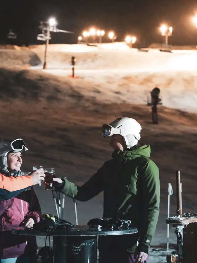 Un groupe de skieurs trinque en terrasse, derrière eux se trouve une piste de ski nocturne éclairée ainsi qu'un télésiège