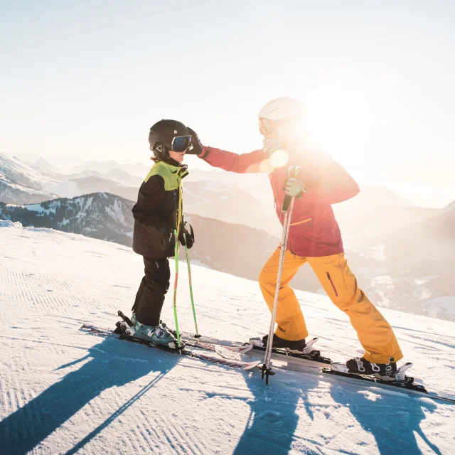 Un parent et son enfant en ski sont sur une piste damé avec en arrière plan la chaine de montagne des aravis enneigée ainsi que le soleil couchant. Le parent replace le masque de ski de son enfant.