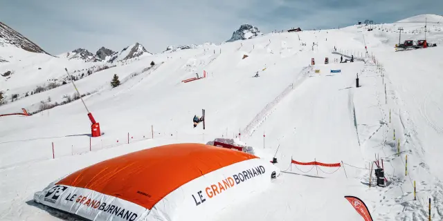 A skier performs a trick in the air before landing on a large orange airbag marked Le Grand-Bornand, in a snowy, mountainous setting.