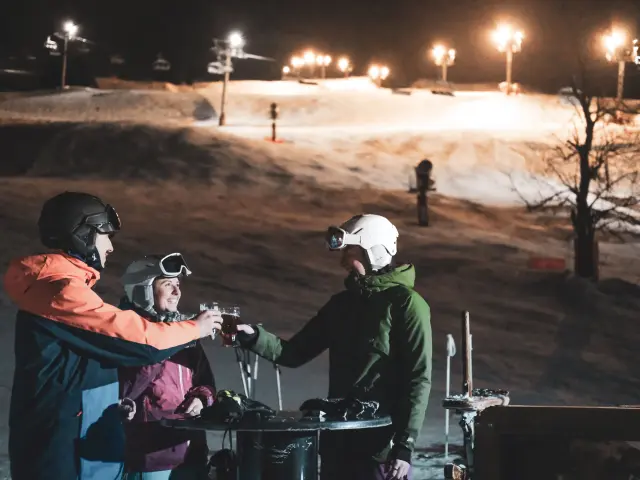 Eine Gruppe von Skifahrern stößt auf der Terrasse an, hinter ihnen befindet sich eine beleuchtete Nachtskipiste sowie ein Sessellift
