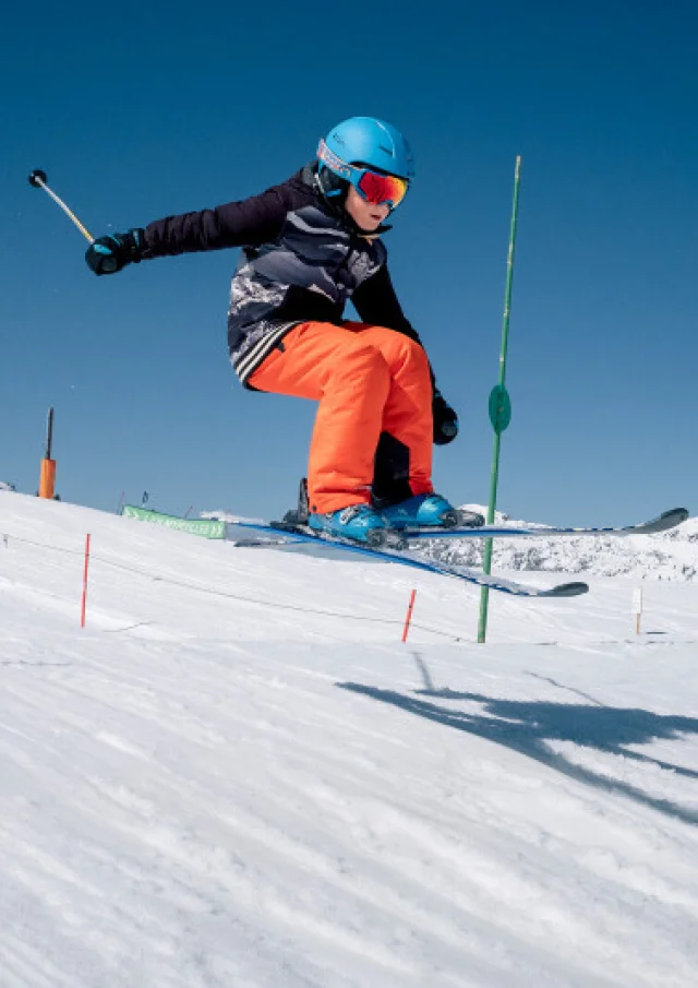 Enfant skieur en plein saut sur une piste enneigée de la Môm'Park au Grand-Bornand, sous un ciel bleu.