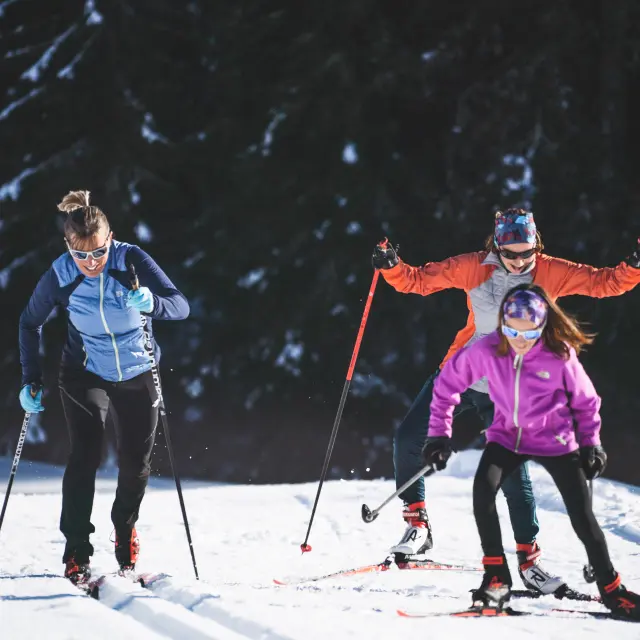 Drei Skifahrer in farbenfroher Kleidung üben Langlauf auf einem verschneiten Weg aus, umgeben von Tannen und Bergen unter einem blauen Himmel.
