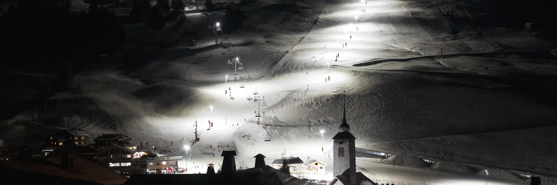 Vue nocturne du domaine skiable du Grand-Bornand illuminé, avec l’église emblématique au premier plan.