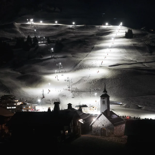 Vue nocturne du domaine skiable du Grand-Bornand illuminé, avec l’église emblématique au premier plan.