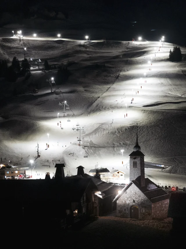 Vue nocturne du domaine skiable du Grand-Bornand illuminé, avec l’église emblématique au premier plan.