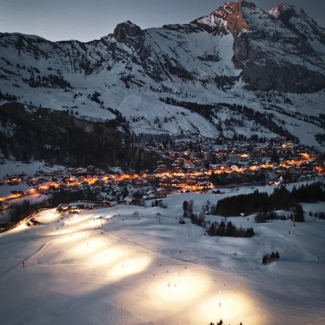 Vertical aerial view of the illuminated Grand Bornand night ski run, with the village of Chinaillon illuminated in the background under the snow-capped mountains of the Bargy massif.