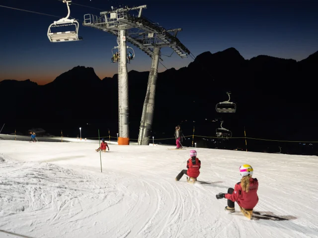 deux skieurs sur des parets descendent la piste de ski nocturne éclairée du grand bornand en passant sous un télésiège pendant qu'en arrière plan on aperçoit les silhouettes nocturne des montagnes
