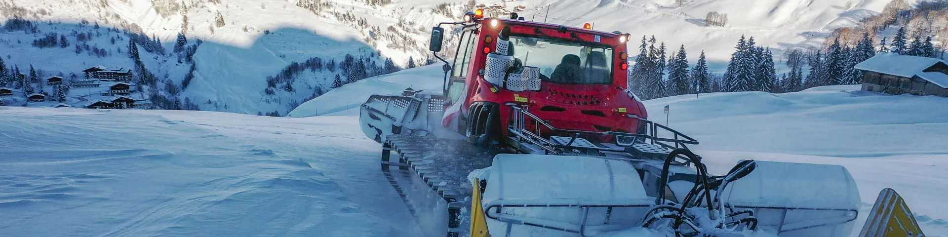 Une dameuse rouge équipée de chenilles et d’une fraise arrière compacte la neige sur une piste de ski, avec en arrière-plan un paysage de montagne enneigé et quelques chalets.