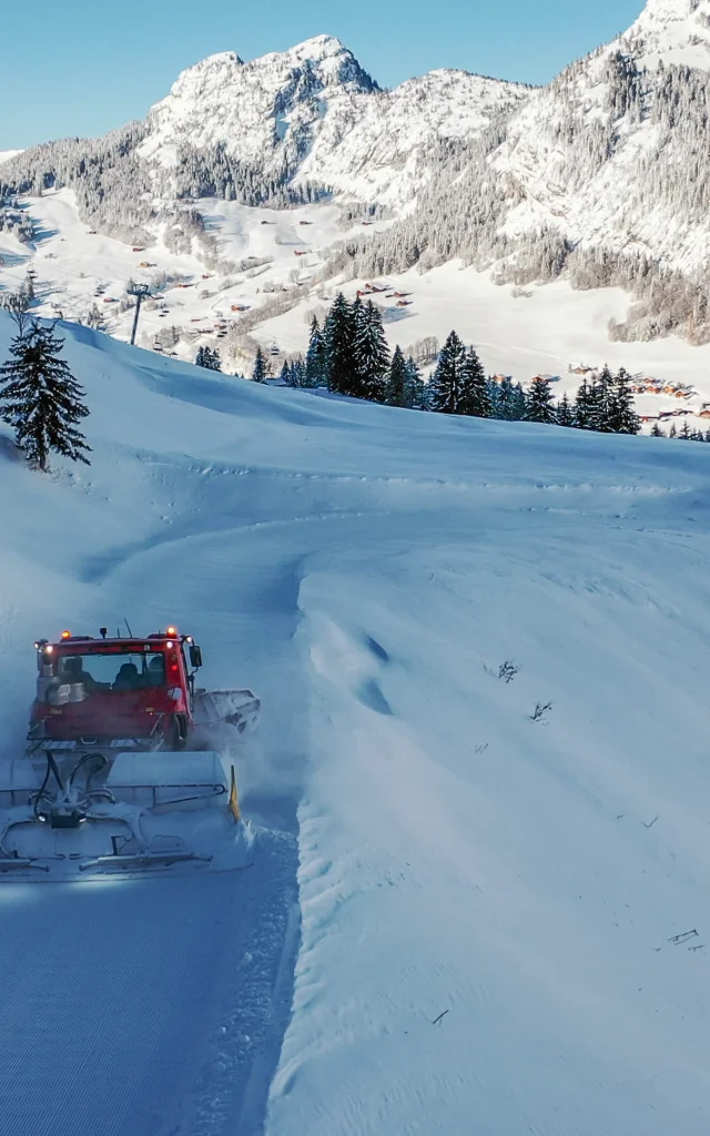 Eine rote Pistenraupe arbeitet auf einer kurvigen, verschneiten Piste, umgeben von schneebedeckten Bergen und Tannen, mit dem Dorf Chinaillon, das unterhalb zu sehen ist.