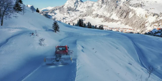 Une dameuse rouge travaille sur une piste enneigée sinueuse, entourée de montagnes enneigées et de sapins, avec le village du chinaillon visible en contrebas.