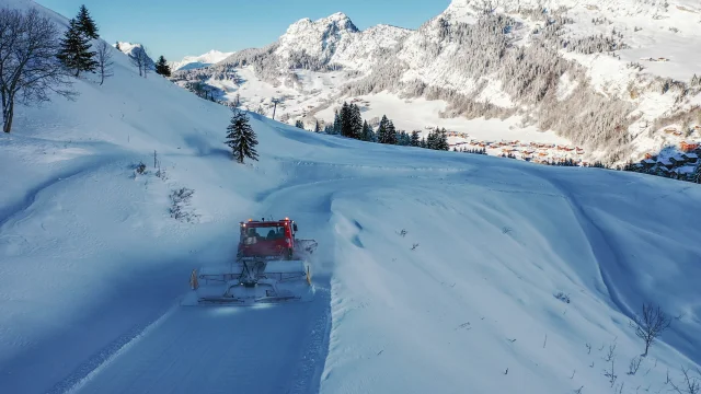 Une dameuse rouge travaille sur une piste enneigée sinueuse, entourée de montagnes enneigées et de sapins, avec le village du chinaillon visible en contrebas.