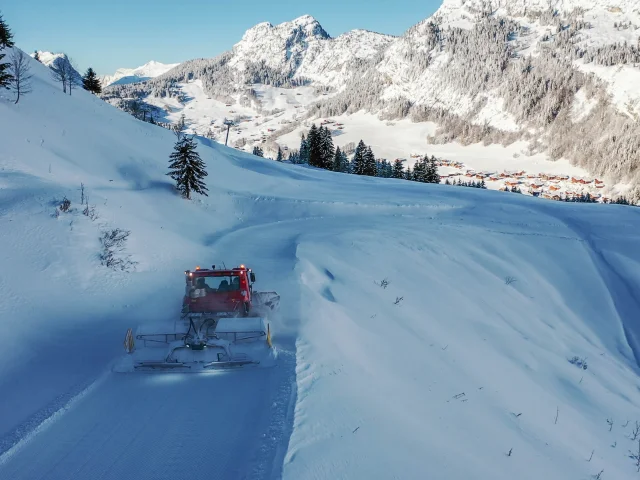 A red groomer works its way down a winding snowy slope, surrounded by snow-capped mountains and fir trees, with the village of Chinaillon visible below.