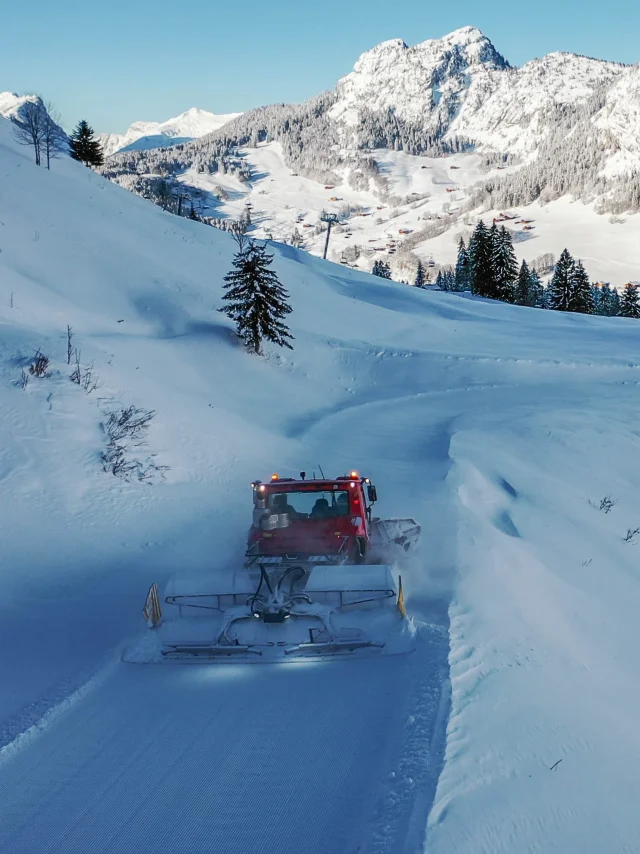Une dameuse rouge travaille sur une piste enneigée sinueuse, entourée de montagnes enneigées et de sapins, avec le village du chinaillon visible en contrebas.